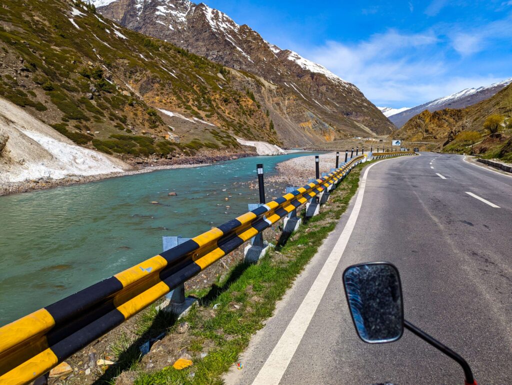 A winding road in Lahaul Valley, flanked by barren landscapes and snow-dusted mountains, with the Chandra River flowing alongside as a constant companion, illustrating the captivating beauty of the journey.