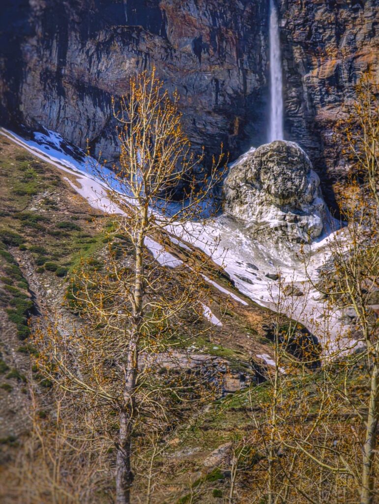 A breathtaking view of the Sissu Waterfall in Himachal Pradesh, cascading from a towering cliff surrounded by snow-covered landscapes and rugged Himalayan look.