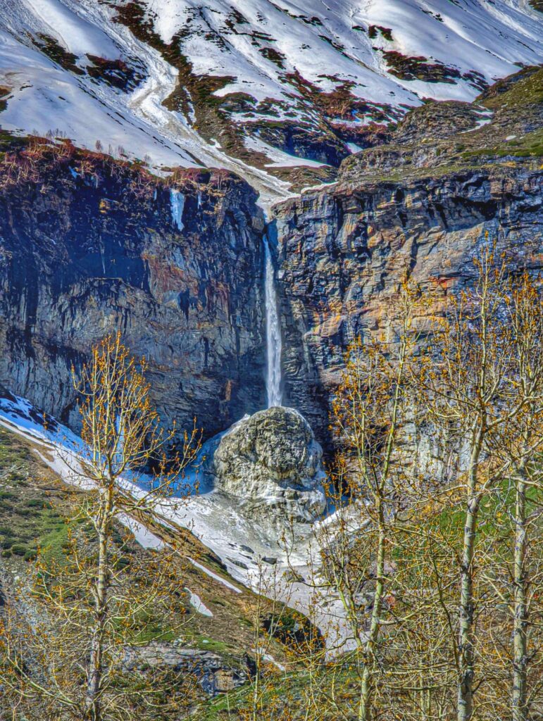 A breathtaking view of the Sissu Waterfall in Himachal Pradesh, cascading from a towering cliff surrounded by snow-covered landscapes and rugged Himalayan look.