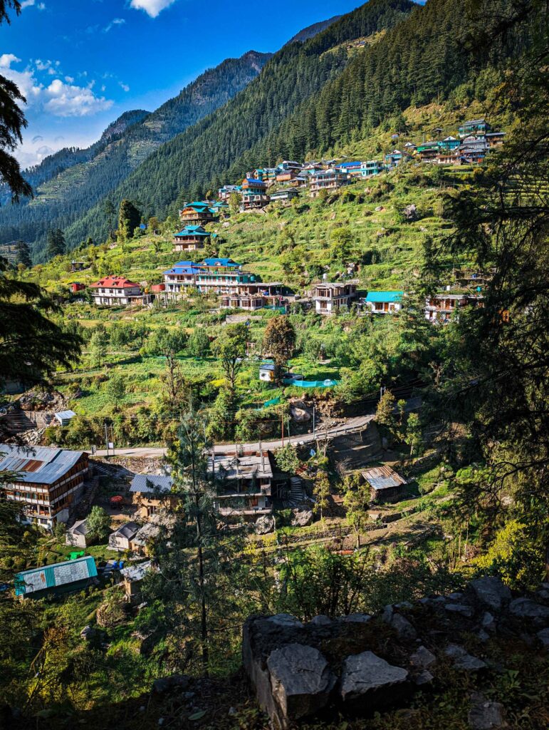 A breathtaking view of Ghiyagi, a charming and peaceful hamlet amidst Himalayas. The scene showcases traditional wooden houses, dense pine forests, and towering snow-capped peaks in the background, capturing the untouched beauty of this hidden village on the scenic route to Shoja."