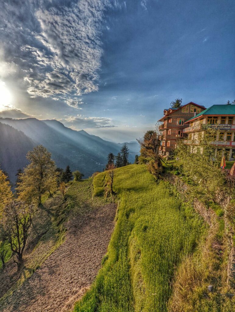 Rustic stays in Shoja, Himachal Pradesh, nestled amidst verdant hills, with a small, well-tended farm in the backyard showcasing rows of fresh vegetables and vibrant flowers. The scene is framed by majestic mountains under a soft afternoon light, creating a peaceful and idyllic atmosphere.