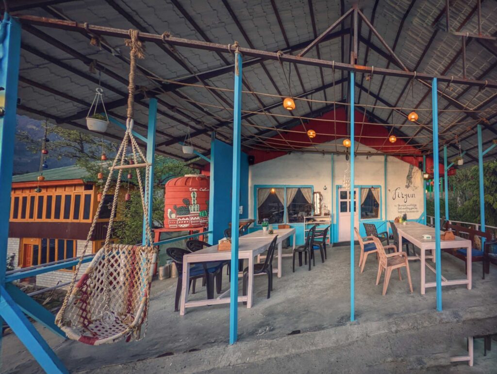Cafe Firgun in Shoja, Himachal Pradesh: A cozy mountain cafe amidst the serene landscapes of the Himalayas. Surrounded by lush greenery, the cafe features wooden interiors, large glass windows offering panoramic views of the valley, and a warm, inviting ambiance perfect for relaxing and enjoying a cup of coffee or a meal.
