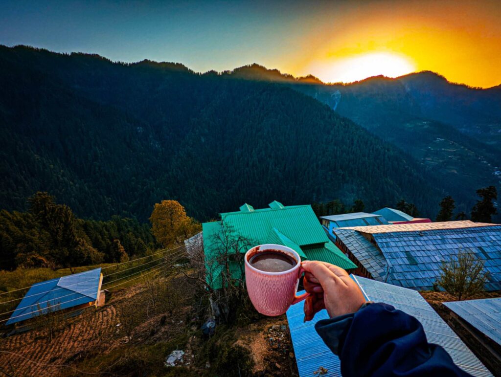 A steaming cup of hot chocolate at Cafe Firgun in Shoja, Himachal Pradesh, enjoyed during a picturesque sunset. The golden hues of the setting sun illuminate the wooden interiors, creating a warm and tranquil atmosphere against the backdrop of the serene Himalayan landscape.
