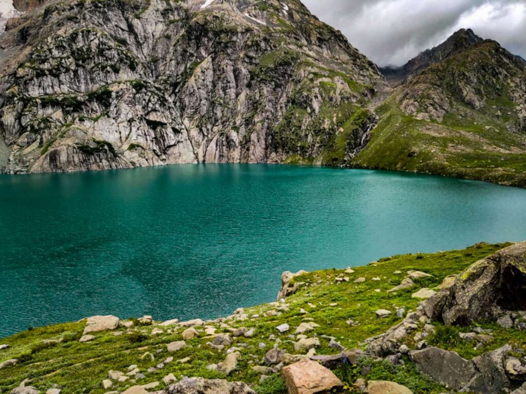 The stunning Gadsar Lake in Kashmir, with its crystal-clear turquoise waters surrounded by lush alpine meadows and towering snow-capped mountains.