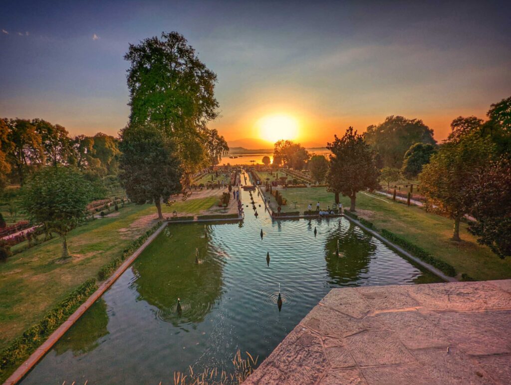A mesmerizing sunset over Dal Lake, viewed from Nishat Bagh in Srinagar, with golden hues reflecting on the serene waters and silhouettes of distant mountains.