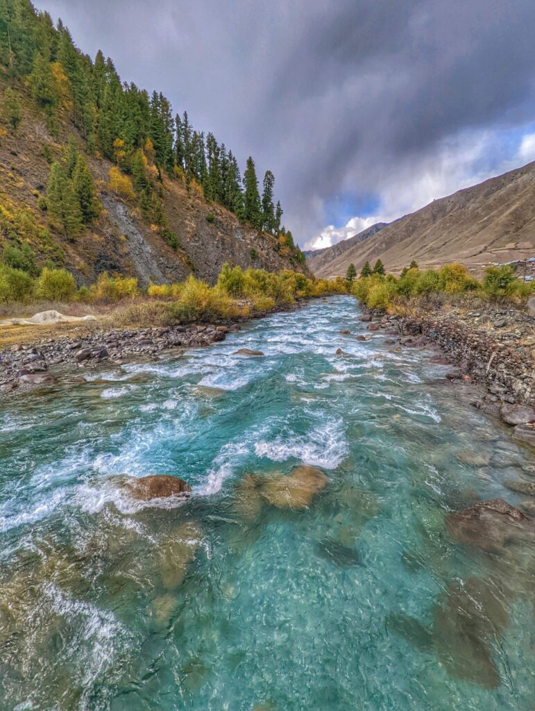 Crystal-clear blue waters of the Kishanganga River winding through rocky banks surrounded by towering pine-covered mountains in the heart of Gurez and Tulail Valley