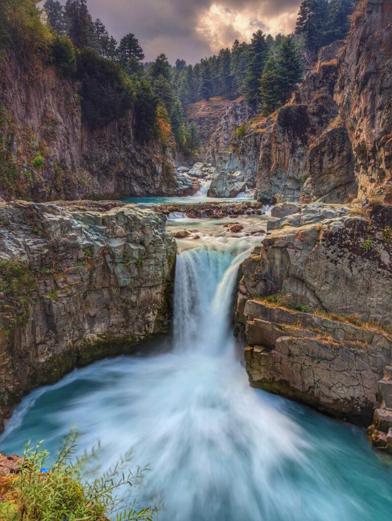 The majestic Aharbal Waterfall in Kashmir, cascading powerfully through rocky terrain, surrounded by lush greenery and a serene atmosphere.