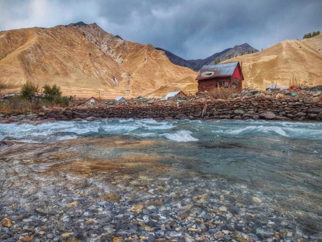 Crystal-clear blue waters of the Kishanganga River winding through rocky banks with a tiny house at backdrop